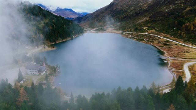 Drone footage of clouds passing over Obersee Lake and the alpine landscape at Staller Sattel. showcases the misty clouds over the lake, surrounded by autumn-colored forests and mountains in the Alps.