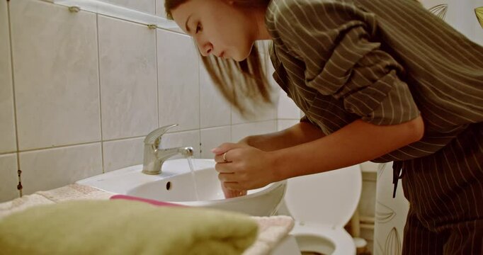 A young woman washes her face in a bathroom sink. She is refreshing herself first thing in the morning.