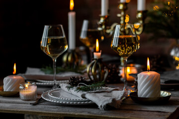 Beautiful table setting for family Christmas dinner at home. Cozy atmosphere, candlelight. Wine glasses, vintage chandelier, elegant interior. Fir tree branches, wooden furniture, dark, bokeh