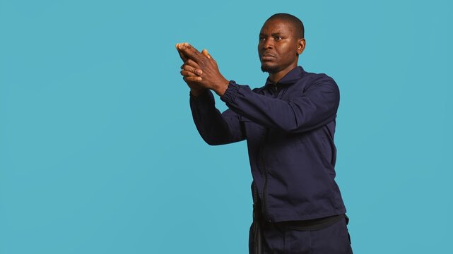 Portrait of security agent securing assigned post, pretending to shoot gun, studio background. African american guardian prepared to handle security breaches, doing pistol fingers gesturing, camera B