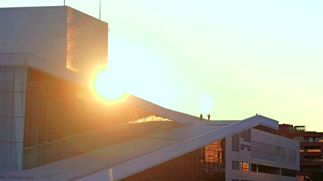 Dramatic sunrise view with silhouettes of people standing on the roof of the Oslo Opera House, capturing the sun&rsquo;s warm glow and architectural details.