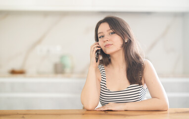 Fototapeta premium Young woman talking on mobile phone sitting at table in kitchen