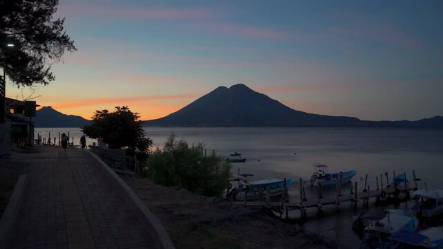 Sunrise on Lake Atitlan, where several boats rest peacefully near a wooden dock. The first rays of the sun illuminate the horizon and reflect in the calm waters of the lake
