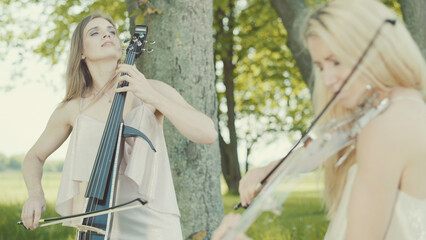 Two beautiful girls in dresses play violin and cello outdoors.