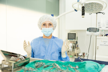 Professional aged female veterinarian in sterile coat, cap, mask and gloves preparing to conduct surgical operation at veterinary clinic, standing by operating table, confidently looking into camera..
