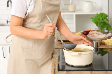 Young housewife with ladle cooking soup on stove in kitchen, closeup