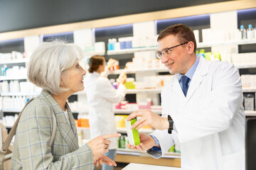 Obraz premium In pharmacy, man apothecary holds package with therapeutic cream for sensitive skin in hands and talks with senior woman client about nuances of care.