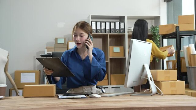 Two young female entrepreneurs run small online clothing store. In a warehouse filled with parcel boxes, they work on laptops, designing clothing and preparing sales brochures and business proposals.