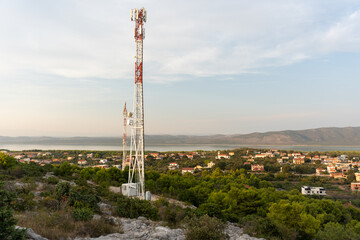 Obraz premium Tall communication towers on rocky hillside overlooking a small village and a scenic coastal landscape with distant mountains and calm water under clear sky. Modern technology and countryside scenery.