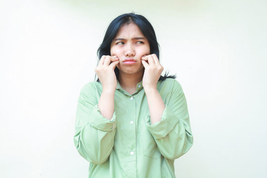 upset asian woman with dark hair touch her cheek wit curled up figer and looking away dissatisfied wearing green oversized shirt isolated on white background
