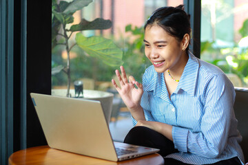 excited asian woman sitting in cafe looking at laptop on wooden table have online meeting, video chat, showing ok signs wearing striped blue shirt