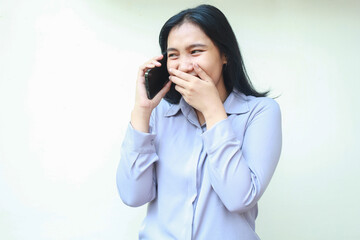 portrait of attractive asian business woman speaking on smartphone covering mouth with hand laughing shy feeling embarrassed looking away standing over isolated white background wear formal shirt