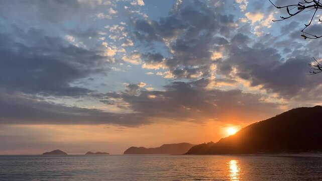 Time-lapse p&ocirc;r do sol. Fim de tarde com o sol se pondo atr&aacute;s de uma montanha e com muitas nuvens no c&eacute;u. Ao fundo, duas ilhas famosas do litoral norte: Ilha das Couves e As Ilhas.