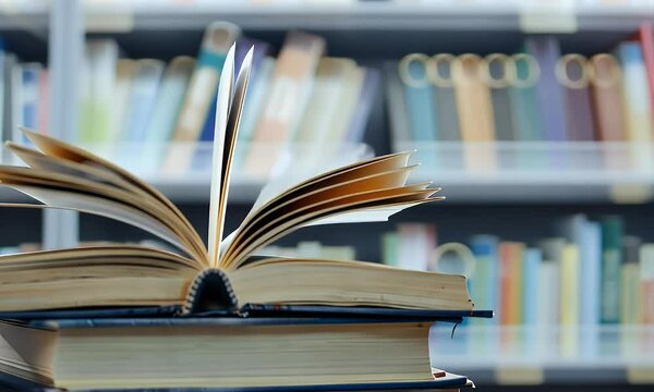 
A stack of books with an open book on top in front of the blurred background of library shelves, symbolizing knowledge and education. 
