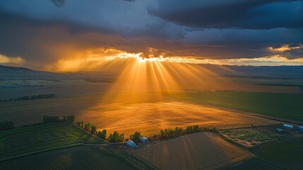 Sunlight streams through storm clouds, illuminating a small patch of farmland with a warm glow, while the surrounding landscape remains under shadow