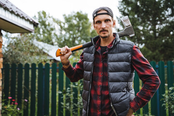 A confident man wearing a flannel shirt and a stylish puffer vest holds an axe in a green garden, surrounded by vibrant nature and greenery © Aleksey
