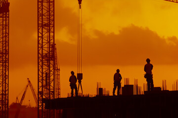 Silhouette of construction workers at sunset, towering cranes against orange sky, urban development in progress