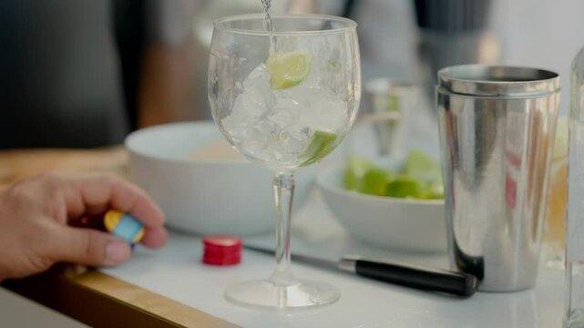 gin and tonic glass filled with ice and lime slices, with a hand holding a bottle in the foreground and a cocktail shaker nearby