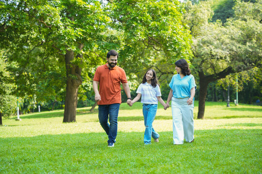 Happy young indian parents with child daughter holding hands and walking in summer park, playful family have fun together outdoor, Holidays and vacations, Kid bonding with mom and dad, togetherness.