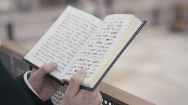 Jewish man praying. He holds a prayer book with Hebrew writing in his hands. Its a close up shot with a background of the Western Wall in Jerusalem, Israel. Action is in slow-motion.