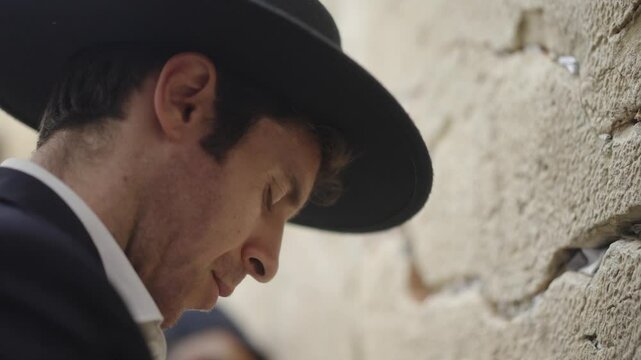 Jewish man praying at the Western Wall in Jerusalem. He wears a black hat and suit. Its a close up shot of his face from side. His eyes are closed as he prays he rocks backward and forwards.