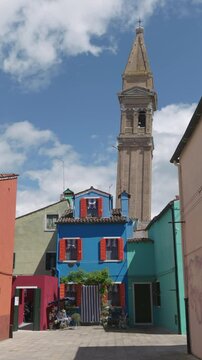 A bright blue house with red shutters stands under the iconic leaning bell tower in Burano, Venice, surrounded by colorful buildings under a partly cloudy sky.