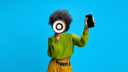 Young African-American woman in green sweater shouting to megaphone and showing phone with blank screen. Copy space. Concept of advertisement, communication, brand promotion, social media, online.