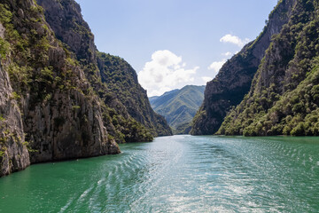 Obraz premium Scenic ferry boat tour on Lake Koman reservoir on Drin river surrounded by forested steep hills, cliffs and majestic rugged mountain ranges of Albanian Alps. Panoramic view along Albanian Fjords. Awe