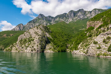 Obraz premium Scenic ferry boat tour on Lake Koman reservoir on Drin river surrounded by forested steep hills, cliffs and majestic rugged mountain ranges of Albanian Alps. Panoramic view along Albanian Fjords. Awe