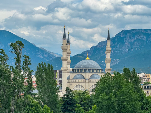 Majestic Namazgah Mosque with twin minarets and a large dome in Tirana, Europe. Surrounded by lush green trees and majestic mountain range Dajti Mountains. Cloudy sky adding dramatic effect to scene