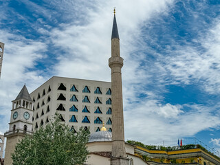 Obraz premium Et'hem Bey Mosque with a tall minaret against a blue sky with white clouds in Tirana, Albania. There is a clock tower next to the mosque, and a modern building with unique facade is visible behind it