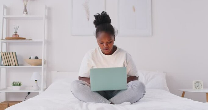 Focused young African American woman sitting on bed with laptop, working or studying from home. Serious black girl deep in thoughts solving problem, thinking about task or planning her next steps.