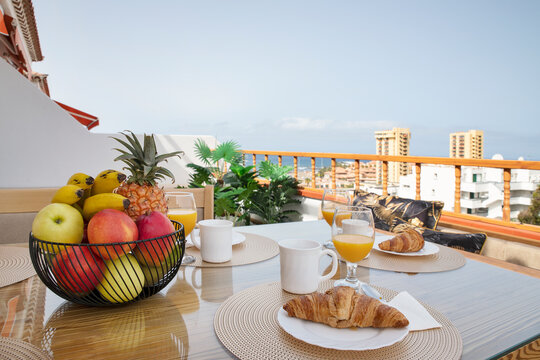 Breakfast with fresh croissants, orange juice and vibrant fruit basket filled with apples, bananas and pineapple set on a high up balcony against the residential views, Tenerife, Canary Islands, Spain
