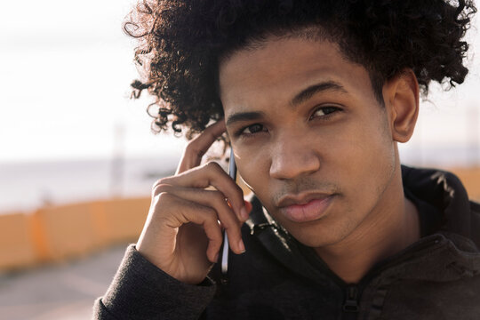 serious african american male teenager talking on smart phone while standing on street looking at camera