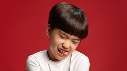 Little boy, Asian child playfully scrunching his face with eyes closed and sticking out tongue against bold red studio background. Concept of facial-expressions, positive and negative emotions. Ad