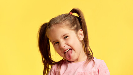 Sticking out her tongue playfully, girl shows off her mischievous side, while keeping lighthearted vibe against sunny yellow background. Concept of facial-expressions, positive and negative emotions.