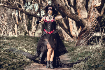 Naklejka premium A young woman in the image of a witch and sorceress, standing in a dark forest, wearing a black dress with a train and a socket on her head.