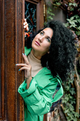 Fototapeta premium Beautiful stylish young woman with curly dark hair posing in a green trench coat in an urban context. Street fashion and style.