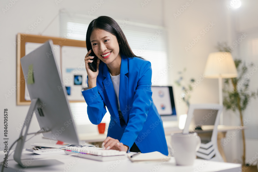 Poster young businesswoman is multitasking at her desk, talking on her phone while working on her computer.