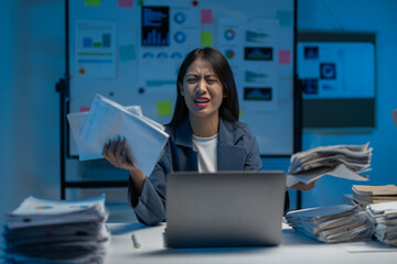 Young businesswoman is holding papers and looking stressed while working late in an office. She is surrounded by paperwork and has a laptop in front of her