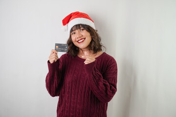 Excited young asian woman wearing santa clause hat and red long sleeved sweater is showing dummy credit card, isolated over white background. Concept for Christmas Holiday and New Year Party