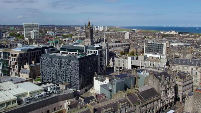 A drone captures the blend of historic and modern architecture in Aberdeen, Scotland, with a clear view of the cityscape and the North Sea coastline. Wind turbines are visible offshore.