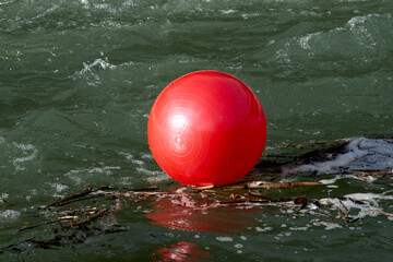 A bright red exercise ball floating on water, caught amidst debris and foam in a fast-flowing river. The ball’s vibrant color contrasts sharply with the dark green water, creating a striking visual.