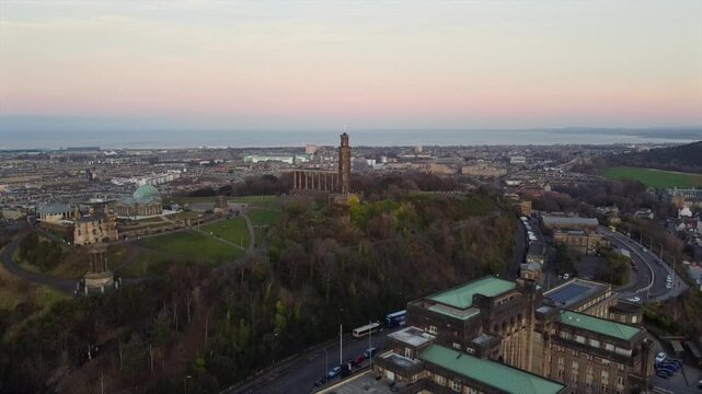Drone footage over Calton Hill in Edinburgh at dawn, featuring historic monuments like the Nelson Monument and National Monument, with a view of the cityscape and coastline in the background.