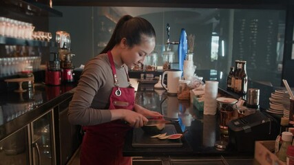 Side view of young Asian barmaid in red apron cutting fruit for cocktails while working in restaurant