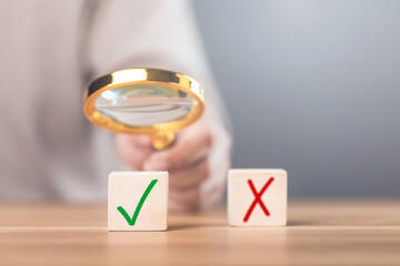 A man holding a magnifying glass over checkmark and 'X' symbols on wooden blocks. The image reflects the concept of choosing right from wrong, highlighting careful decision-making and evaluation