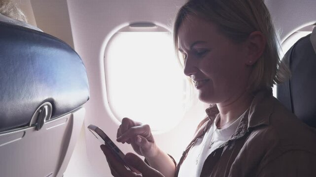 Passenger woman is flying in plane. Girl using phone sitting by airplane window. Traveling female with smartphone inside plane enjoying flight. Woman using wifi connection on board. Traveling girl