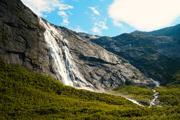 Obraz premium A beautiful view of sunlit glacial waterfalls falling from great heights into a forest stream on a warm, sunny day in Briksdal, Norway
