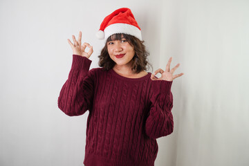 Excited young asian woman wearing santa clause hat and red long sleeved sweater is showing OK Hand Gestures, isolated over white background. Concept for Christmas Holiday and New Year Party