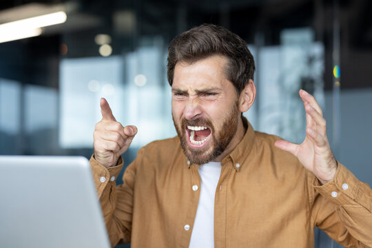 Frustrated businessman yelling at laptop during video call. Intense anger and stress evident in facial expressions. Office setting hints at workplace conflict, emotion, frustration, and tension.